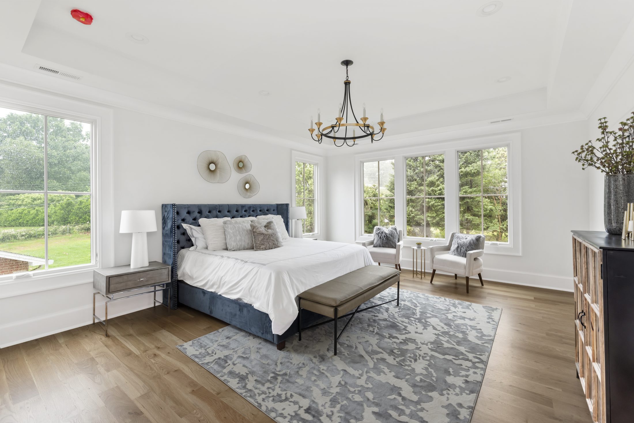 Primary bedroom with blue tufted bed, tray ceiling, iron chandelier, and a window-bay sitting area — Bywood by Stillman Properties Charlotte NC