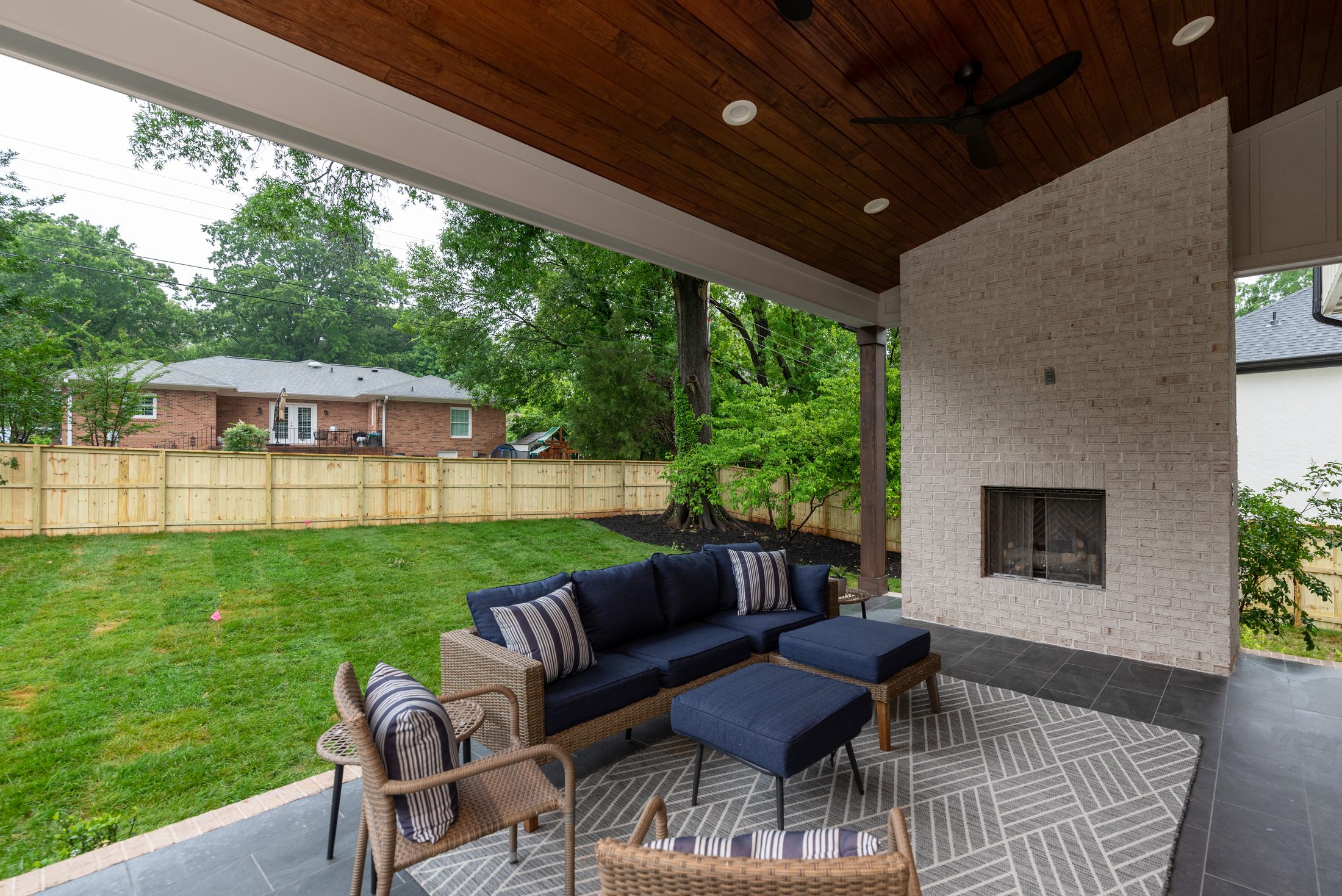 Covered outdoor living room with cedar ceiling, modern ceiling fan, and whitewashed brick fireplace overlooking the backyard — Bywood by Stillman Properties Charlotte NC
