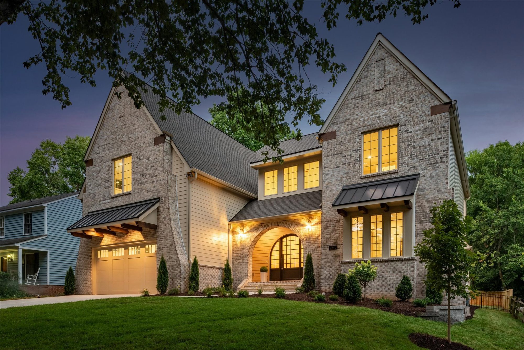 Heather custom home in Charlotte NC by Stillman Properties — brick and lap-siding exterior at twilight with steep twin gables, standing-seam metal awnings, and an arched entry aglow