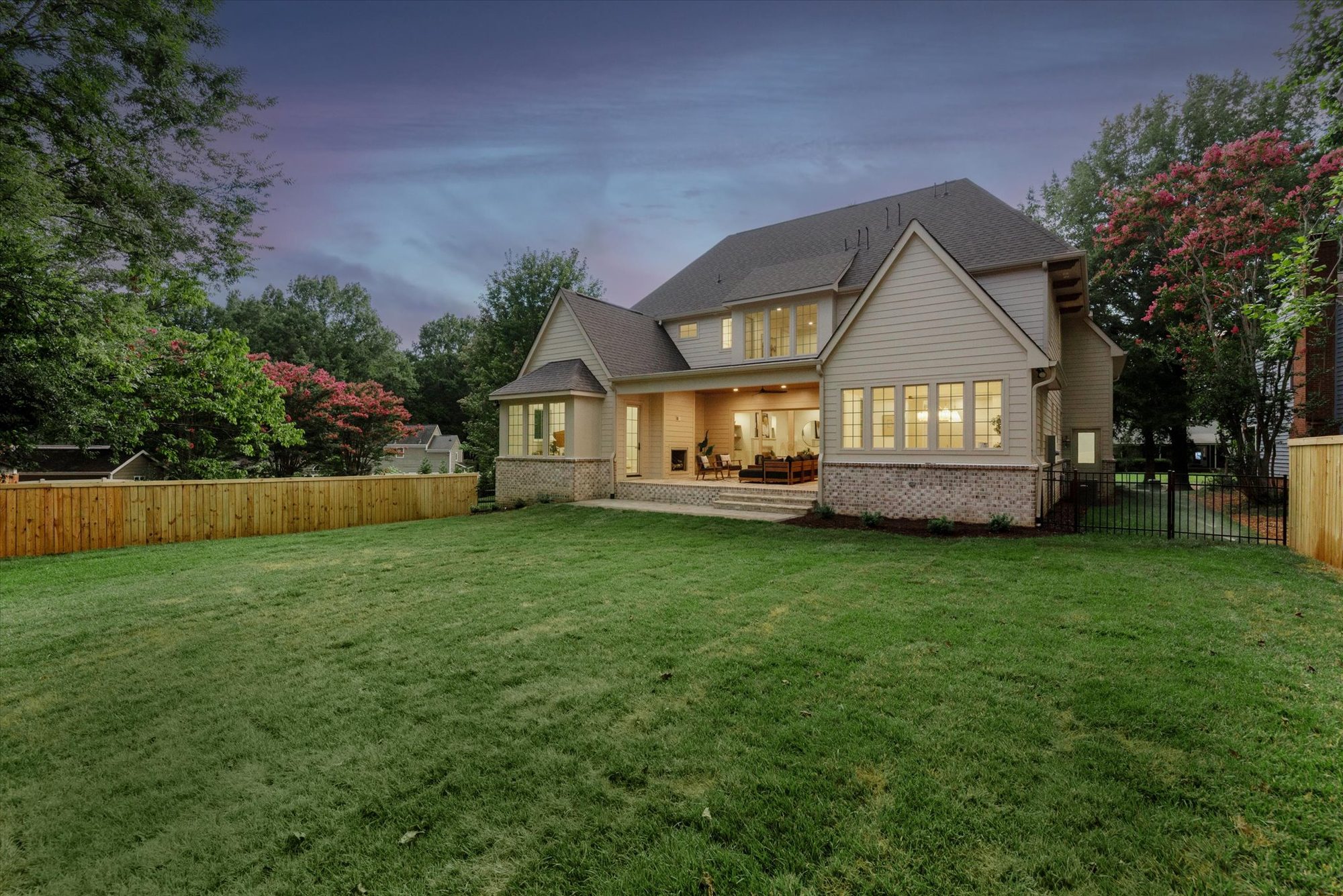 Rear elevation of Heather at twilight with the covered porch and great room windows aglow above the fenced lawn — Heather by Stillman Properties Charlotte NC